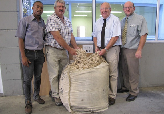 Four individuals standing behind a burlap sack filled with mohair cuttings, placed in front of a small wall sign featuring the words 