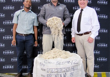 Three individuals standing behind a large bale of wool, with a backdrop featuring the logo 