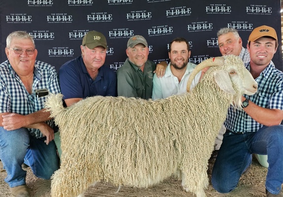 Group of six individuals posing with a large sheep at an event, standing in front of a backdrop with the logo of The House of Fibre.